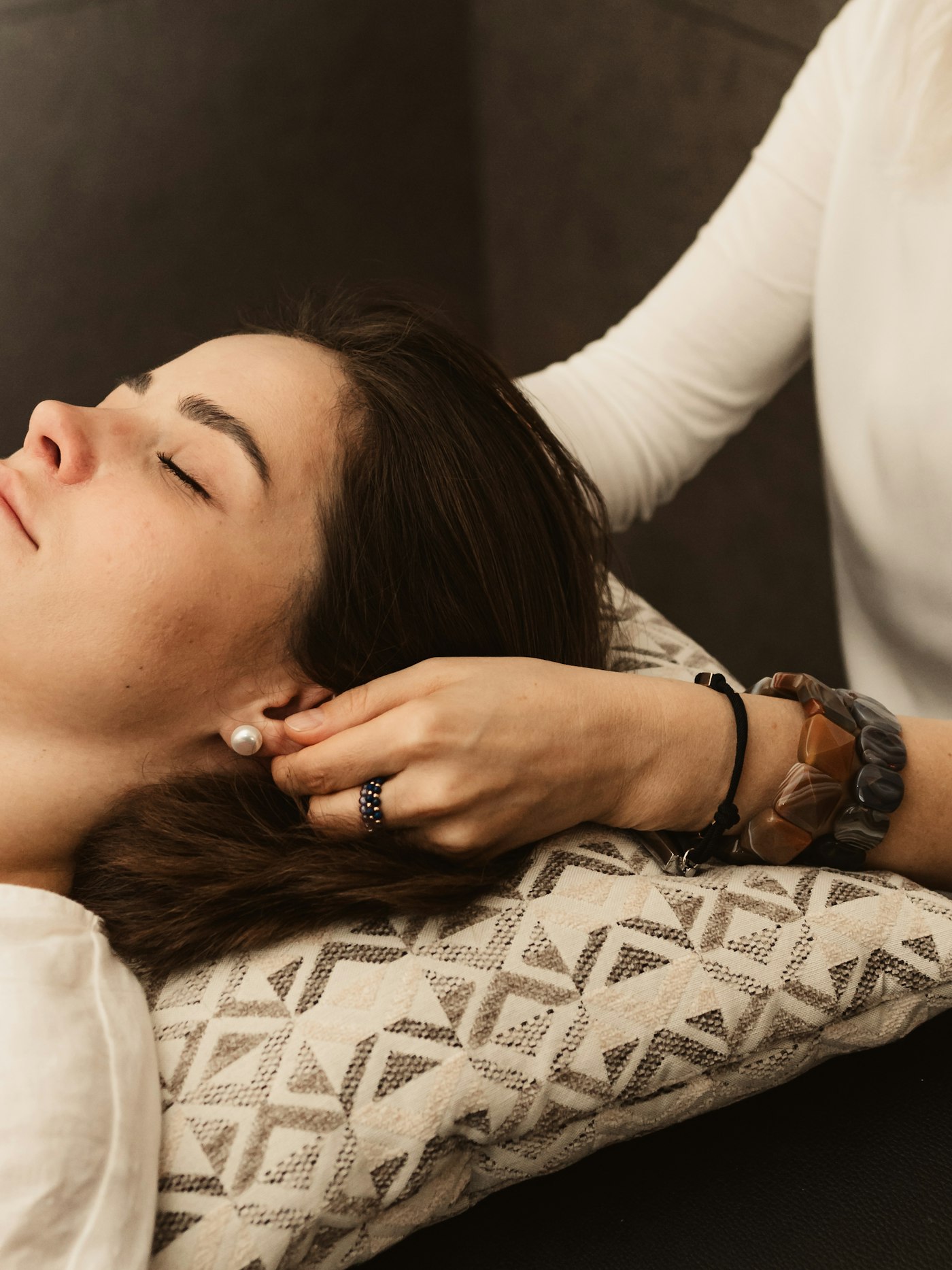 Close view of a guest lying with her eyes closed on a woven cream cushion while a therapist cradles the base of her skull with one hand