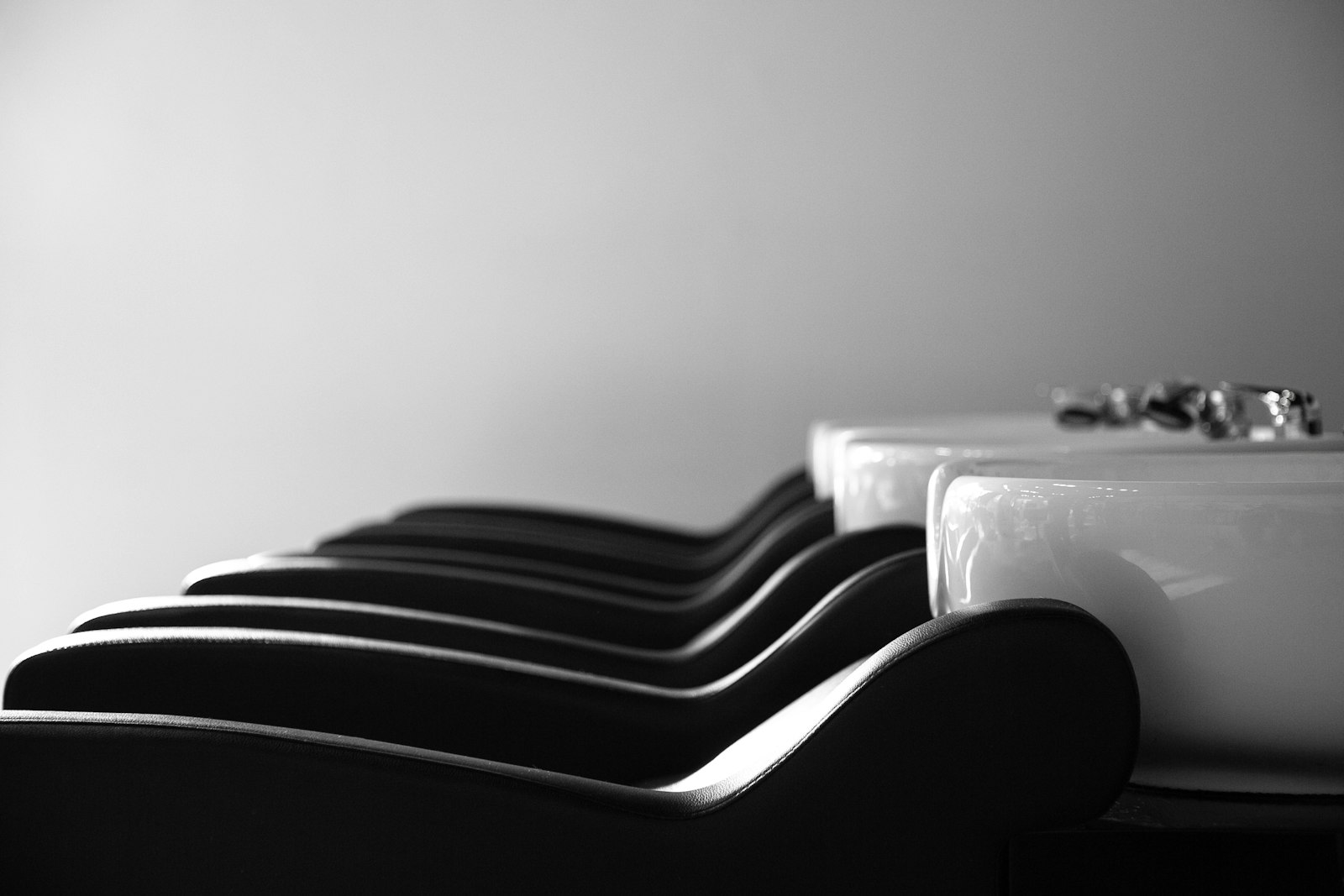 Black and white photograph of four black reclining chair headrests in a row with white ceramic basins behind them, against a plain pale wall