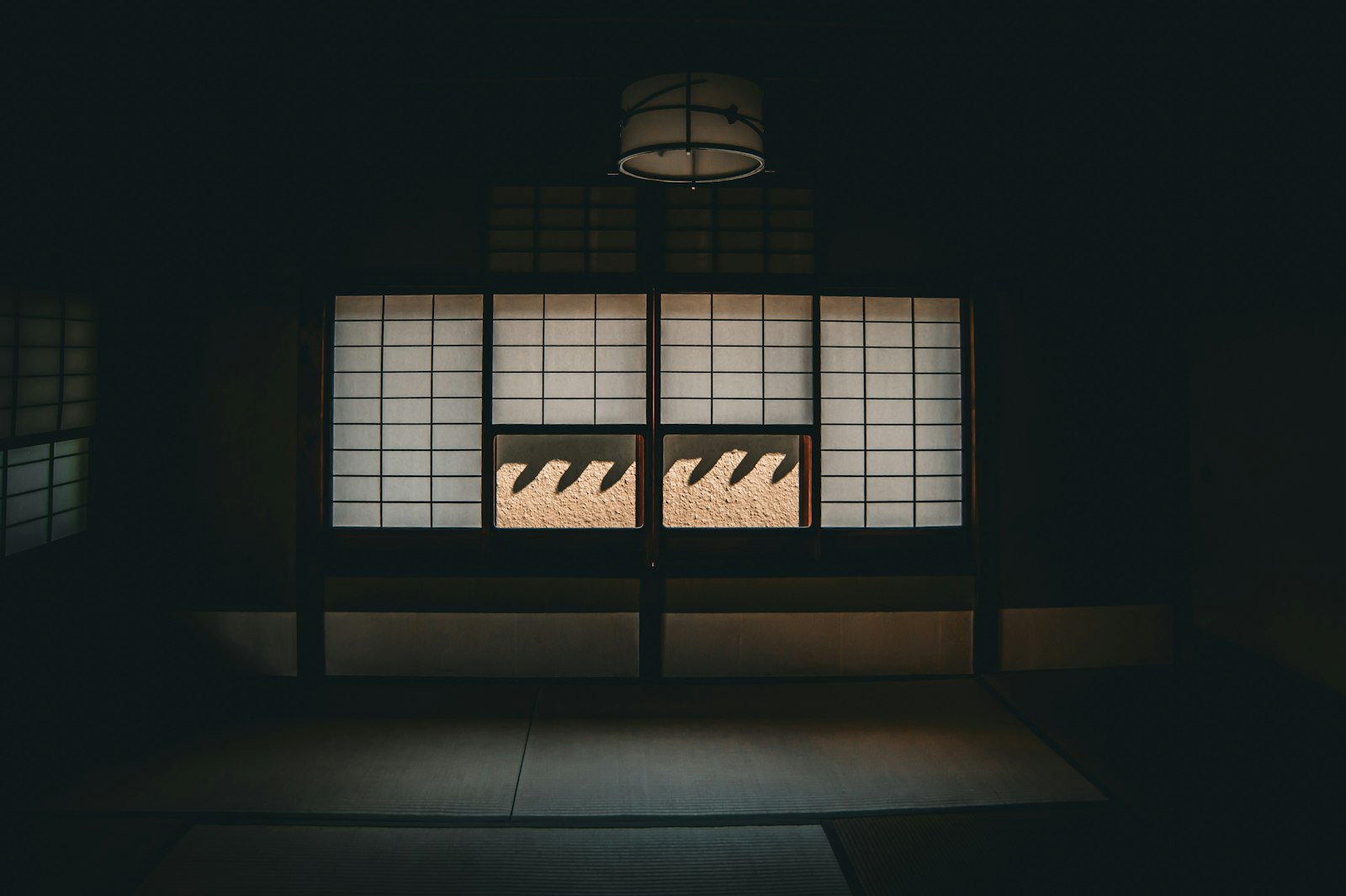 A dim room seen head on, with a wall of glowing white paper screens, two lower panels open onto a sunlit wall, and a cylindrical paper lantern above